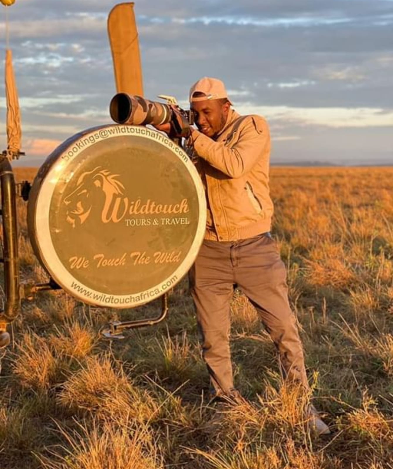 Jacob Munene of Wildtouch Tours & Travel beside the Wildtouch brand emblem at golden hour in the East African bush